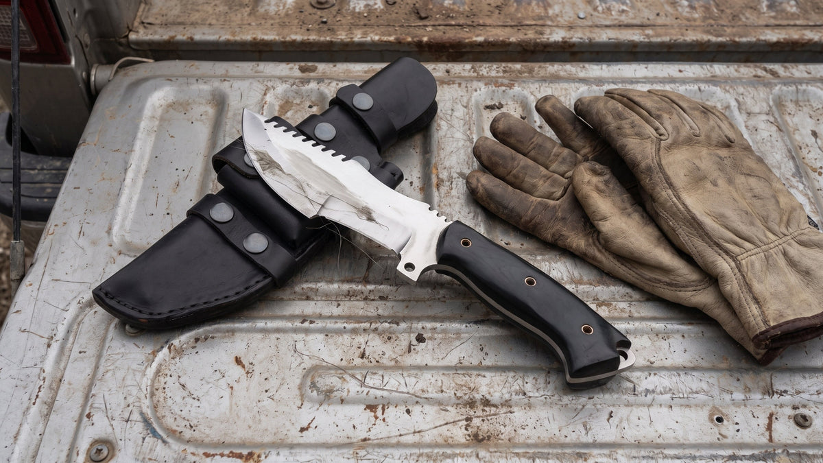 Tracker D2 full tang knife and its black sheath on the tailgate of a pickup truck beside work gloves