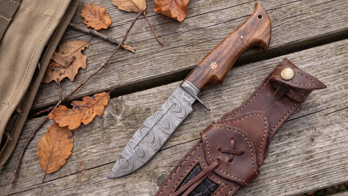 Beast Hunter Damascus hunting knife and its leather sheath on a weathered picnic table at an autumn hunting camp