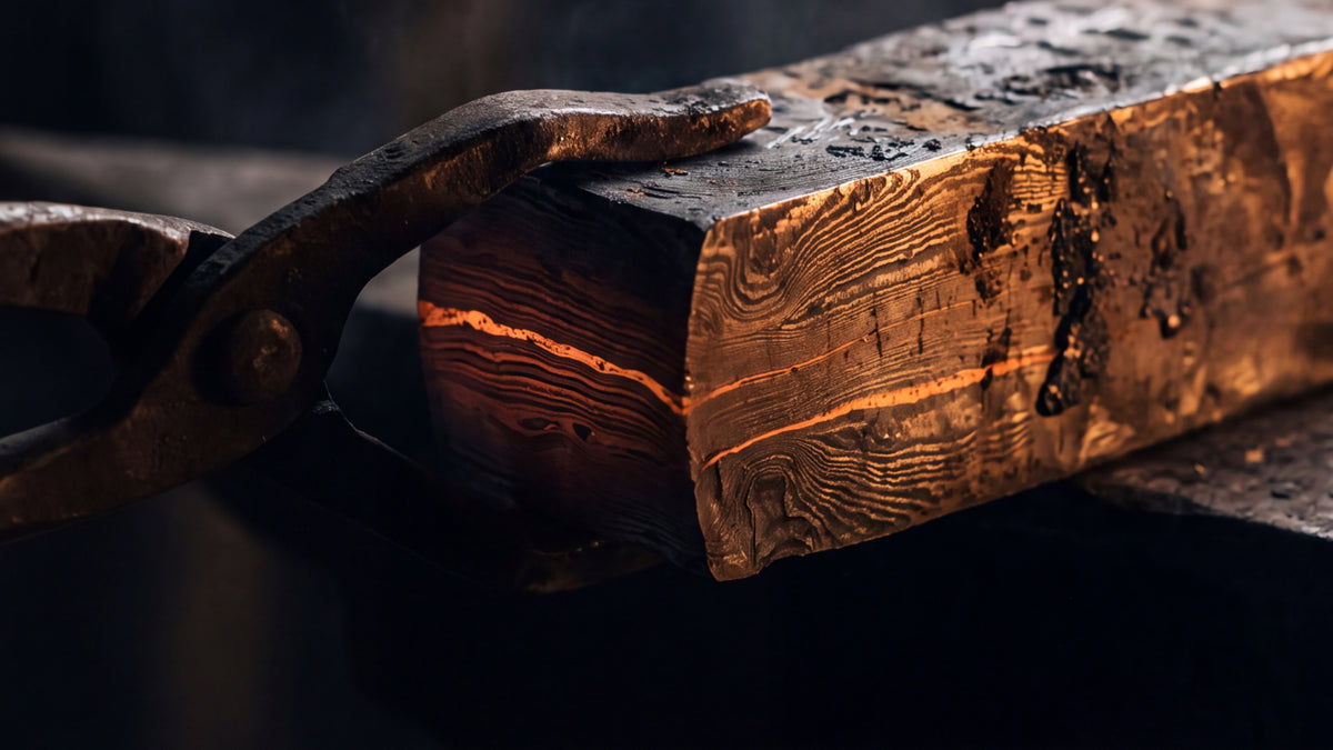 Tongs gripping a layered steel billet, the folded layers showing on the cut end