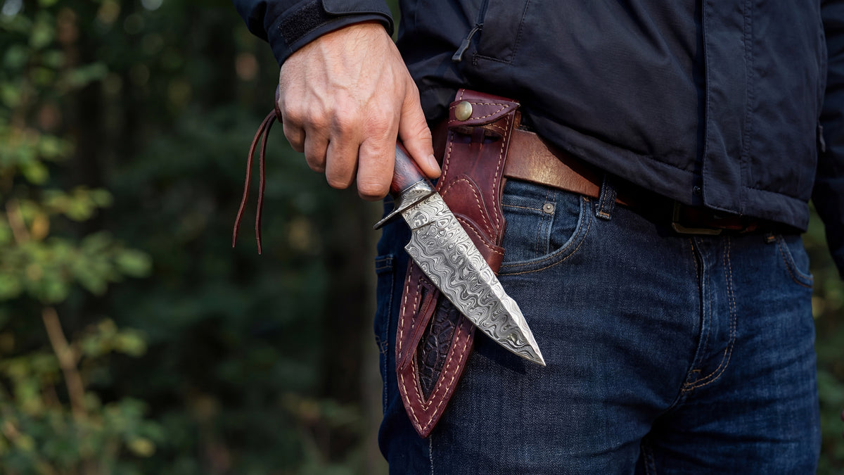 Drawing a Damascus knife from a brown leather sheath worn on a belt, woodland behind